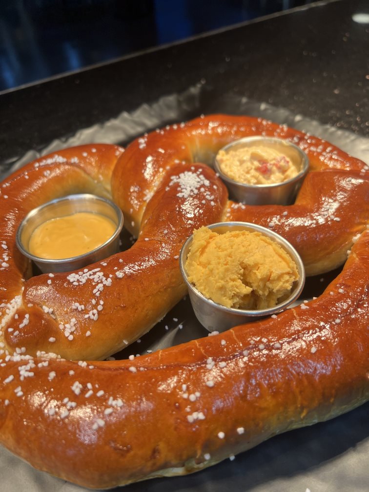 A large pretzels sprinkled with coarse salt. In the pockets sit dip containers filled with pub, pimento, and hot beer cheese.