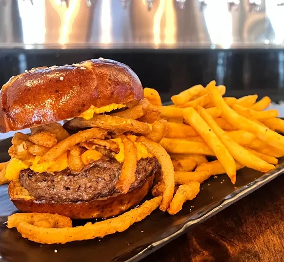 A hamburger on a pretzel bun. It is topped with a pile of fried onion rings and a yellow pub spread. There is a side of french fries next to it.