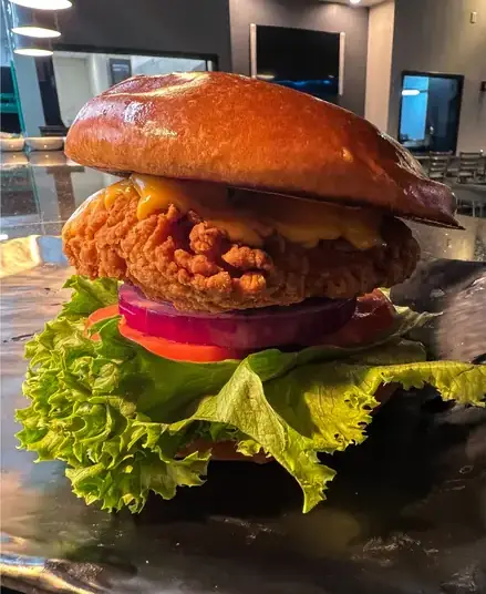 A fried chicken sandwich. The chicken patty is sitting atop a tomato, large greens, and an onion.