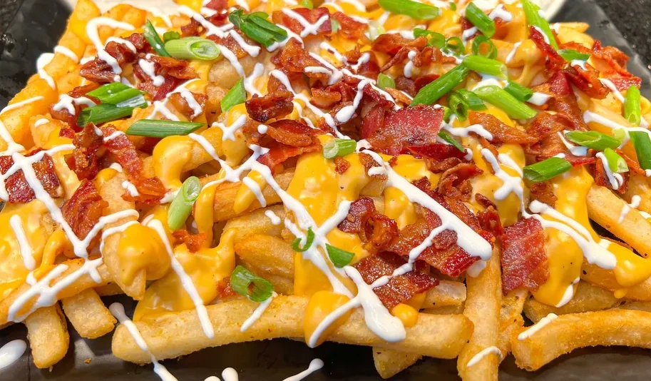 A plate of french fries covered with melted cheese, crumbled bacon, chives, drizzled in a crisscross pattern.