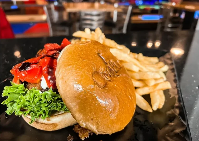 A sandwich with the bun off to the side. Atop a fried and marinated mushroom cap are lettuce greens, bright red tomatoes, and goat cheese. The bun top is branded with the On Par logo. In the background is a side of fries.