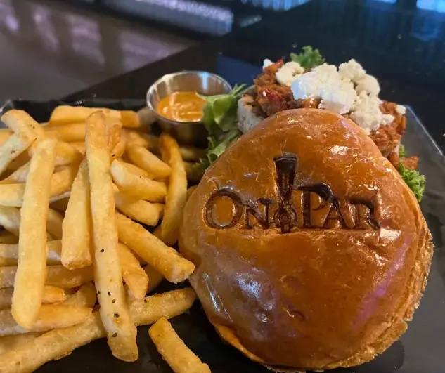 A plate with a hamburger with the On Par logo branded on the top alongside a side of fries and cup of dip. The hamburger is covered with pesto and goat cheese.